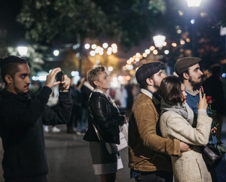 Zdjęcie z nocnego mapingu na budynku Dużej Sceny Teatru. Ujęcie przedstawia planty koło budynku Teatru. Kilka osób stoi z telefonami komórkowymi w rękach, rejestrując pokaz mapingu.