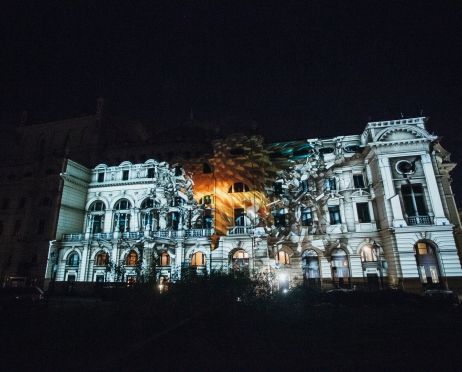 Zdjęcie z nocnego mappingu na budynku Dużej Sceny Teatru. Ujęcie obejmuje cały gmach od strony plant. Mapping ukazuje w centralnym miejscu bocznej ściany ogień oraz osuwające się ściany Teatru.