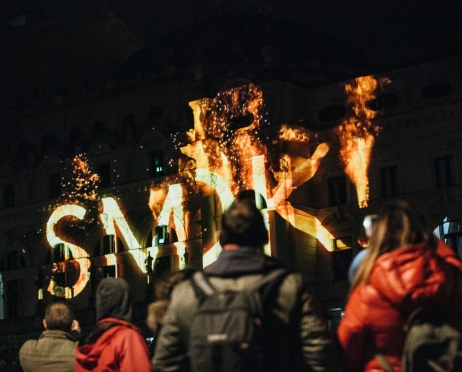 Zdjęcie z nocnego mapingu na budynku Dużej Sceny Teatru. Ujęcie obejmuje fragment gmachu od strony plant, na którym wyświetlono napis ”SMOK”. Napis robi wrażenie jakby płonął – czcionka jest w kolorze ognia, za nim są płomienie. Miejscami napis jest zasłaniany przez ludzi oglądający pokaz.