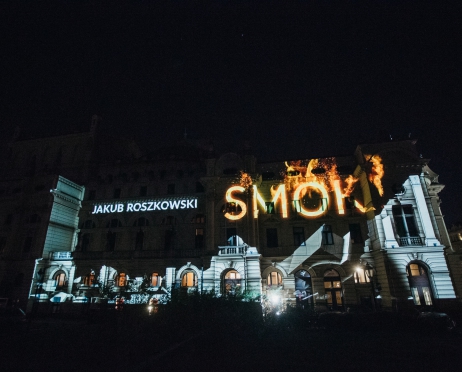 Zdjęcie z nocnego mapingu na budynku Dużej Sceny Teatru. Ujęcie obejmuje cały gmach od strony plant. Wyświetlono na nim nazwisko autora i reżysera - „JAKUB ROSZKOWSKI” oraz tytuł „SMOK”. Napis smok sprawia wrażenie jakby płonął – czcionka jest w kolorze ognia, za nim są płomienie