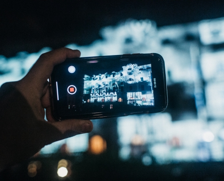 Na zdjęciu trzymany bokiem telefon komórkowy, nagrywający film. Na ekranie smartfona boczna ściana budynku Dużej Sceny Teatru od strony plant, oświetlona na niebiesko. W tle zamazane kontury budynku.