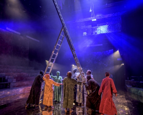 Na zdjęciu grupa postaci w różnokolorowych strojach ustawiająca wielką drabinę. Dookoła półokrągłe ławy jak w antycznym teatrze.