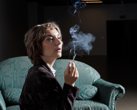 Zdjęcie przedstawia portret kobiety. Siedzi bokiem. Patrzy przed siebie. Jest zamyślona. W ręku trzyma palącego papierosa. Dym unosi się w górę. Ubrana jest w brązowo białą garsonkę. Za nią zielona kanapa. Tło jest czarne.