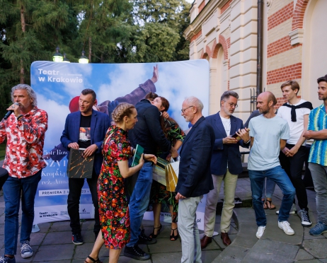 Na zdjęciu rozproszona grupa osób. Po lewej stronie mężczyzna w średnim wieku w ręku trzyma mikrofon coś mówi. Ubrany jest w kolorową koszulę i jeansy. Obok niego grupa osób, która jest odwrócona bokiem. Wyglądają jakby szli przed siebie. W ręku mają torebki prezentowe. W tle zielone drzewa.