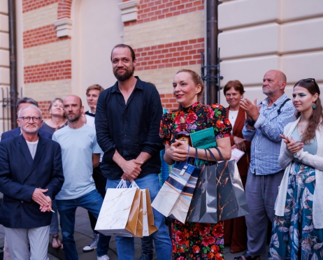 Zdjęcie przedstawia taras. W centralnej części na środku kobieta i mężczyzna w średnim wieku. Uśmiechają się patrzą przed siebie. W dłoni trzymają torby prezentowe. Kobieta ubrana jest w kolorową sukienkę w kwiaty, mężczyzna w czarną koszulę i niebieskie spodnie. Za nimi grupa osób. Patrzą w ich kierunku. Tło jest kremowe.