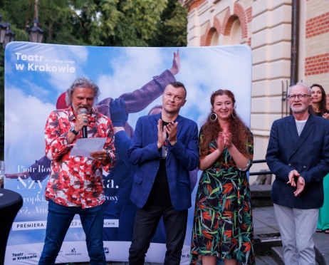 Na zdjęciu w centralnej części po lewej stronie mężczyzna. Czyta coś z kartki, ma mikrofon. Ubrany jest w kolorowa koszulę i jeansy. Obok niego mężczyzna w średnim wieku, uśmiecha się i klaszcze. Ubrany jest w niebieską marynarkę i czarne spodnie. Po prawej stronie uśmiechnięta kobieta. Klaszcze. Ubrana jest w kolorową sukienkę w kwiaty. W tle ścianka materiałowa w kolorze niebieskim.