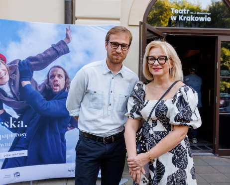 Na zdjęciu w centralnej części kobieta i mężczyzna w średnim wieku. Pozują na ściance centralnej przed teatrem. Uśmiechają się. Kobieta ubrana jest w biało czarną sukienkę. Ma blond włosy. Mężczyna ma białą koszulę i czarne spodnie. W tle szklane drzwi do sali teatralnej.