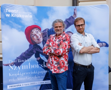 Na zdjęciu w centralnej części dwóch mężczyzn w średnim wieku. Pozują na ściance teatralnej. Ręce mają skrzyżowane na wysokości klatki piersiowej. Uśmiechają się. Mężczyzna po lewej stronie ubrany jest w kolorową koszulę oraz jeansy. Mężczyzna po prawej ma białą koszulę i czarne spodnie.