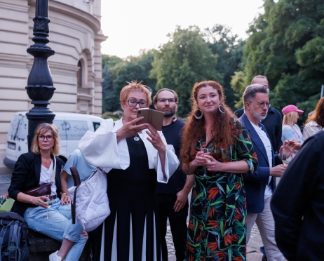 Na zdjęciu grupa ludzi. Parzą w jednym kierunku. Słuchają kogoś. Jedna z kobiet trzyma  w ręku telefon, robi zdjęcia. W tle budynek teatru.