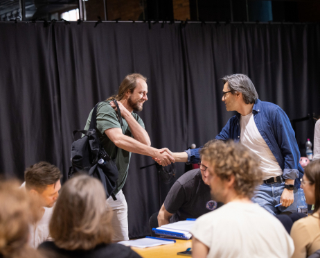 W sali prób Teatru przy ulicy Radziwiłłowskiej, na tle czarnej kotary, 35-letni aktor Antoni Milancej i scenarzysta Robert Jarosz witają się uściskiem dłoni. Antoni Milancej ma długie jasnobrązowe włosy związane z tyłu, krótką brodę i wąsy. Ubrany jest w zielony T-shirt i jasne spodenki, a na ramieniu ma przewieszony czarny plecak. Naprzeciw niego stoi Robert Jarosz,  mężczyzna o siwiejących włosach zaczesanych na bok, w okularach, ubrany w białą koszulkę i rozpiętą niebieską jeansową koszulę. Mężczyźni uśmiechają się podczas powitania. Na pierwszym planie, przy długim stole, siedzą pozostali uczestnicy próby, widoczni głównie od tyłu.