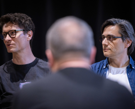 W sali prób Teatru przy ulicy Radziwiłłowskiej obok siebie siedzą 48-letni reżyser Paweł Chomczyk i 49-letni scenarzysta Robert Jarosz podczas próby czytanej spektaklu „Układ Scalony”. Po lewej stronie Paweł Chomczyk ma krótkie ciemne włosy zaczesane na bok, jasną cerę i czarne okulary w grubych oprawkach. Ubrany jest w ciemnoszarą koszulkę z kolorowym nadrukiem. Po prawej stronie Robert Jarosz ma średniej długości, siwiejące włosy zaczesane na bok, okulary w ciemnych oprawkach oraz niebieską jeansową koszulę narzuconą na biały T-shirt. . Na pierwszym planie, nieostro, widoczna jest sylwetka jednego z uczestników próby, częściowo zasłaniająca obu twórców. W tle znajduje się ciemna przestrzeń sali prób.