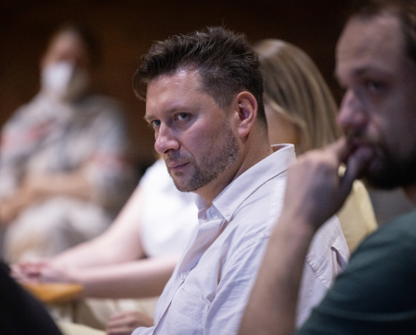 W sali prób Teatru przy ulicy Radziwiłłowskiej przy stole siedzi przedstawiciel Centrum Nauki Cogiteon. To mężczyzna o krótkich, ciemnych włosach zaczesanych do góry, z krótką brodą i wąsami oraz jasną cerą. Ubrany jest w białą koszulę z długim rękawem. Patrzy uważnie przed siebie z poważnym, skupionym wyrazem twarzy, przysłuchując się dyskusji podczas próby czytanej spektaklu. Po bokach kadru, lekko nieostro, widoczni są pozostali uczestnicy spotkania, a w tle znajduje się ceglana ściana charakterystyczna dla industrialnej przestrzeni sali prób.