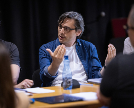 W sali prób Teatru przy ulicy Radziwiłłowskiej 49-letni scenarzysta Robert Jarosz siedzi przy stole podczas próby czytanej spektaklu „Układ Scalony”. Ma średniej długości, siwiejące włosy zaczesane na bok, okulary w ciemnych oprawkach i ubrany jest w niebieską jeansową koszulę narzuconą na biały T-shirt. Gestykuluje obiema dłońmi, zwracając się do pozostałych uczestników spotkania. Na stole przed nim leżą egzemplarze scenariusza, laptop i butelka z wodą. Wokół niego, częściowo nieostro, siedzą członkowie zespołu realizatorskiego i obsady spektaklu.
