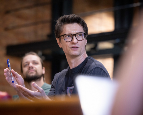 W sali prób Teatru przy ulicy Radziwiłłowskiej 48-letni reżyser Paweł Chomczyk siedzi przy stole podczas próby czytanej spektaklu. Ma krótkie, ciemne włosy zaczesane na bok, jasną cerę, szczupłą twarz i nosi czarne okulary w grubych oprawkach. Ubrany jest w ciemnoszarą koszulkę z kolorowym nadrukiem. W prawej dłoni trzyma niebieski długopis, a otwartymi dłońmi żywo gestykuluje. W tle, lekko nieostro, siedzi 35-letni aktor Antoni Milancej. Ma długie ciemne włosy związane z tyłu, krótki zarost i ubrany jest w zielony T-shirt. Z uniesioną głową przysłuchuje się rozmowie. W tle widoczne są ceglane ściany i industrialna przestrzeń sali prób.