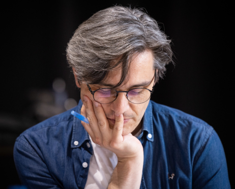W sali prób Teatru przy ulicy Radziwiłłowskiej scenarzysta spektaklu „Układ Scalony” Robert Jarosz siedzi pochylony nad scenariuszem. To mężczyzna o siwiejących, gęstych włosach zaczesanych na bok, z pojedynczym pasmem opadającym na czoło. Ma na sobie białą koszulkę i rozpiętą niebieską jeansową koszulę oraz okrągłe okulary w cienkich, ciemnych oprawkach. Opiera lewy policzek na dłoni, między palcami trzyma niebieski długopis. Tło zdjęcia jest ciemne.