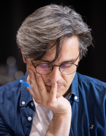 W sali prób Teatru przy ulicy Radziwiłłowskiej scenarzysta spektaklu „Układ Scalony” Robert Jarosz siedzi pochylony nad scenariuszem. To mężczyzna o siwiejących, gęstych włosach zaczesanych na bok, z pojedynczym pasmem opadającym na czoło. Ma na sobie białą koszulkę i rozpiętą niebieską jeansową koszulę oraz okrągłe okulary w cienkich, ciemnych oprawkach. Opiera lewy policzek na dłoni, między palcami trzyma niebieski długopis. Tło zdjęcia jest ciemne.