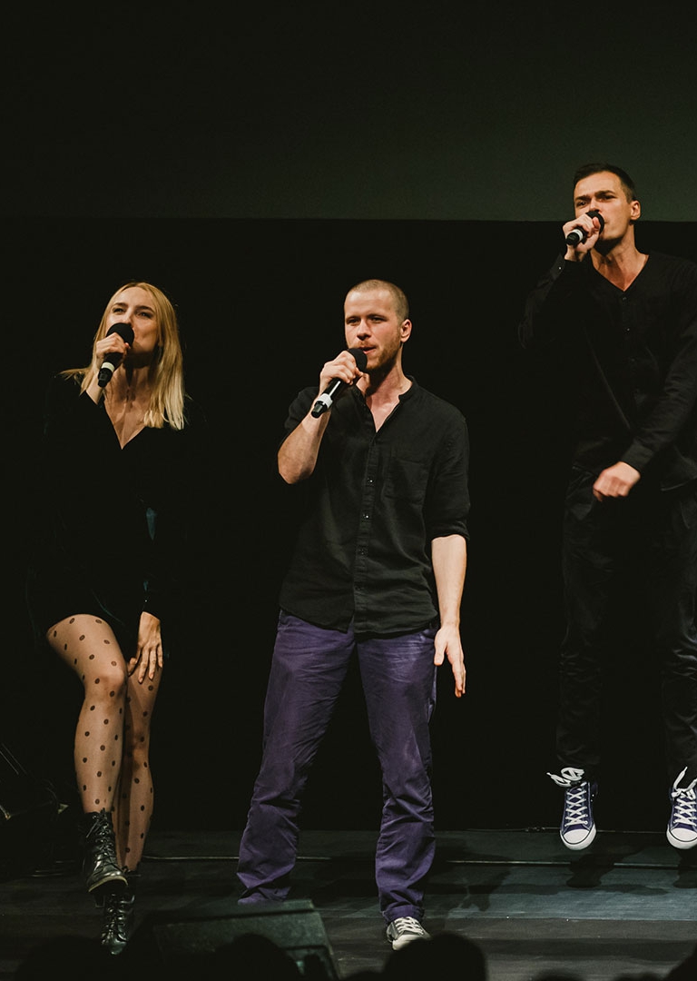 Na zdjęciu aktorzy Teatru, od lewej: Daniel Malchar, Karolina Kazoń, Rafał Szumera. Trzymają przy ustach mikrofony, śpiewają. Aktor z prawej podskoczył i unosi się w powietrzu. Mają ciemne, współczesne ubrania.