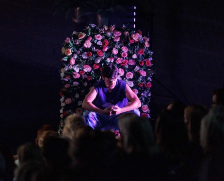 Ciemne zdjęcie. Na środku konstrukcja fragmentu ściany, cała obłożona różowymi kwiatami. Przed nią siedzi mężczyzna ubrany na fioletowo. Dłonie ma złączone, ma opuszczoną głowę w dół. Na pierwszym planie cienie głów osób siedzących na widowni.