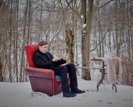 Mężczyzna siedzi w czerwonym fotelu, w dłoniach trzyma dwie różdżki do szukania żył wodnych. Jest ubrany na czarno ale na głowie ma małą, czerwoną czapkę - toczek. Przed nim stoi mały, okrągły stoliczek. Za nim rośnie mały las. Na ziemi leży śnieg.