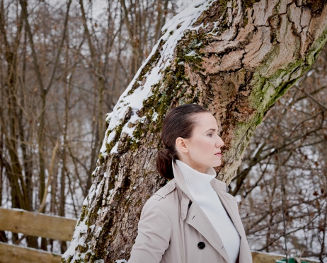 Na zdjęciu kobieta opiera się bokiem o drzewo. Ma włosy związane w kucyk. Ubrana jest w jasny płaszcz i biały golf. Patrzy przed siebie. W tle zaśnieżony las.