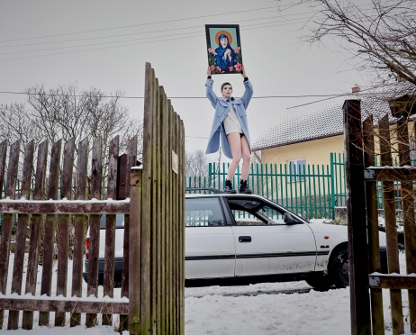 Na zdjęciu dziewczyna w niebieskim płaszczu stoi na dachu białego samochodu. Unosi ręce do góry. Trzyma w nich obraz Maryi. Samochód stoi na drodzę. Widać tylko część, bo zasłania go drewniany płot. Na ziemi leży śnieg.