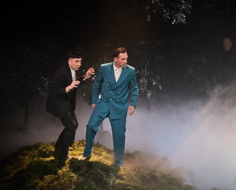 Na zdjęciu dwóch mężczyzn stoi na górce zrobionej ze sztucznej trawy. Jeden z nich ma na sobie niebieski a drugi czarny garnitur. Wyglądają, jakby przed czymś uciekali. Tło jest czarne. Przy ziemi unosi się dym.