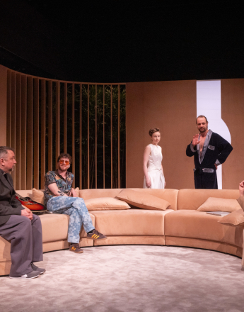 Scena MOS Teatru Słowackiego. Na pierwszym planie widoczna jest rozbudowana, półokrągła sofa w odcieniach beżu, ustawiona na jasnej, miękkiej wykładzinie. Minimalistyczna scenografia utrzymana jest w ciepłych, piaskowych tonach, tło tworzą zaokrąglone ściany oraz pionowe drewniane lamele, za którymi widać roślinność. Po bokach sceny znajdują się wysokie, białe podświetlane elementy dekoracyjne przypominające abstrakcyjne formy architektoniczne. Od lewej stoi Šimon Krupa jako Honza Kovács ubrany w jasny, kremowy sweter i szare spodnie. Na sofie siedzi Marek Daniel jako Zdeněk Dvořák w ciemnoszarej marynarce, szerokich szarych spodniach i sportowych butach. Obok niego Daniel Malchar jako Natan ma na sobie kolorową koszulę we wzory, jasno niebieskie spodnie w kratę oraz okulary z pomarańczowymi szkłami. W głębi sceny stoi Kateřina Měchurová jako KOMENSKÝ 2.0 w roli ubrana w białą, prostą sukienkę bez rękawów. Obok niej Antoni Milancej jako Tomek Wielicki ubrany w ciemny, aksamitny szlafrok z połyskującymi wykończeniami, odsłaniającym klatkę piersiową. Po prawej stronie siedzi Anna Tomaszewska jako Bożena Klimek w eleganckim, beżowym garniturze i plisowanej spódnicy, gestykulując uniesionym palcem, jest w trakcie wypowiedzi.