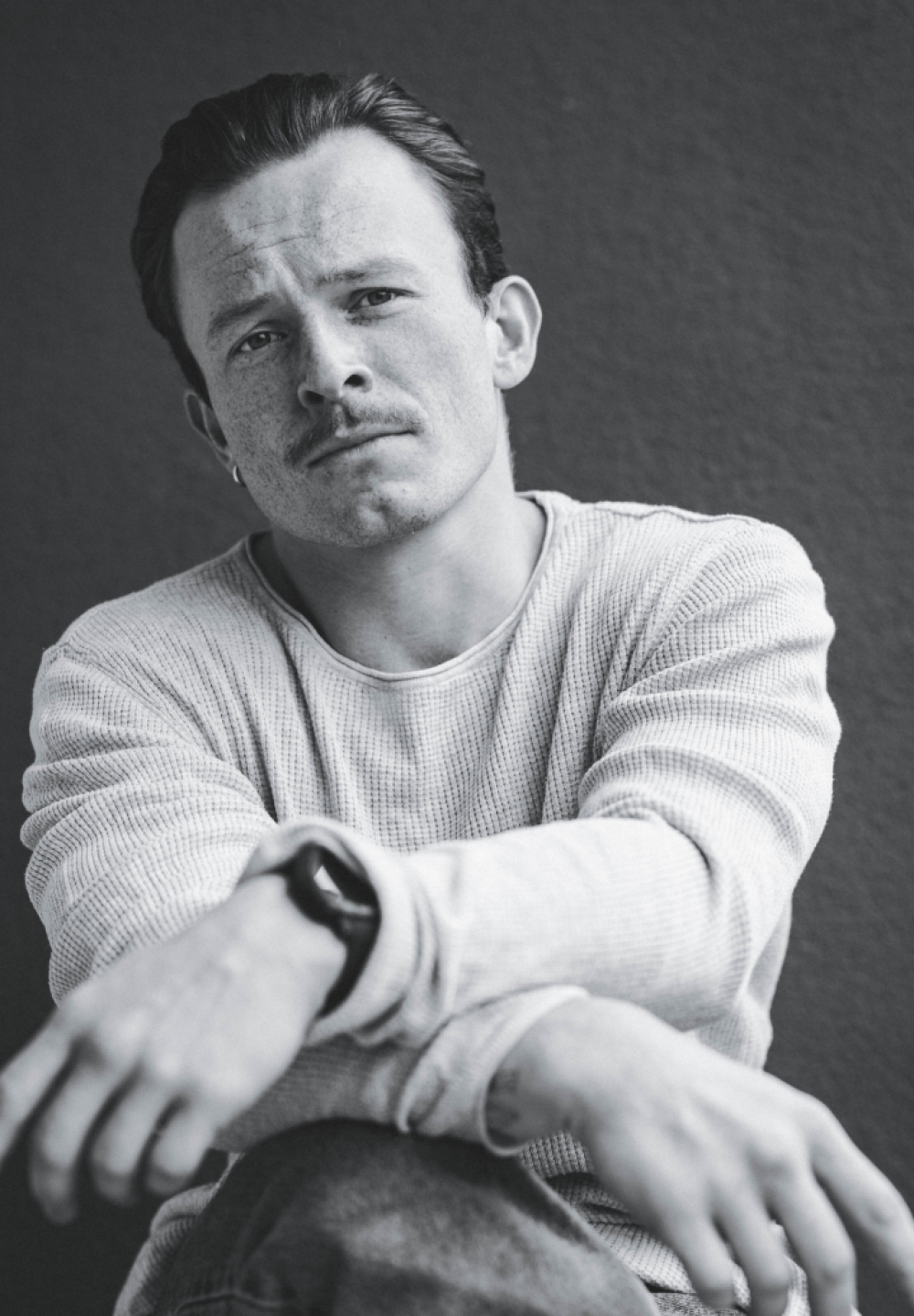 Czarno-biały portret 30-letniego Eryka Pawłowskiego siedzącego w swobodnej, lekko pochylonej pozie. Ma krótkie, zaczesane do tyłu włosy, jasną cerę z widocznymi piegami oraz delikatny zarost i wąsy. Patrzy w obiektyw z poważnym, spokojnym wyrazem twarzy. Ubrany jest w jasny, długorękawowy sweter o wyraźnej fakturze. Na lewym nadgarstku ma zegarek, a w jednym uchu niewielki kolczyk. Jego ręce są wysunięte do przodu, lekko rozmyte przez perspektywę. Tło jest jednolite i ciemne.