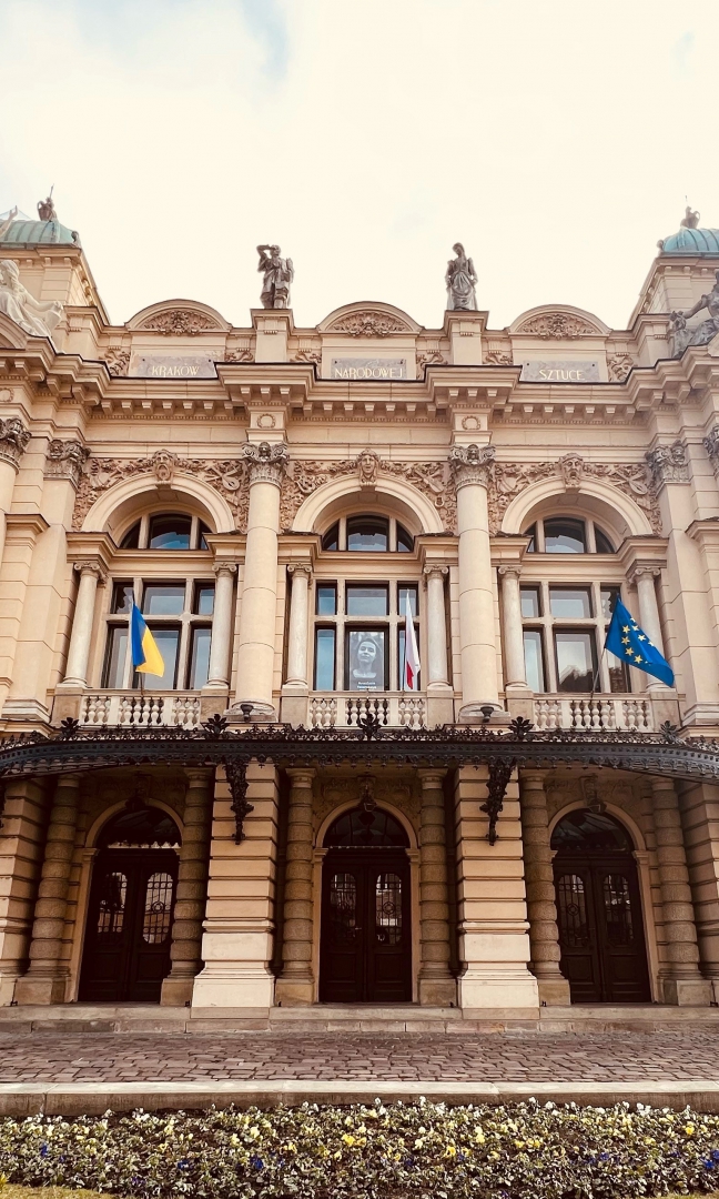 Na zdjęciu w centralnej części front głównego budynku Teatru. Na pierwszym planie wejście główne. Duże masywne ciemne drzwi. Nad drzwiami balkon. Na balkonie zawieszone trzy flagi. Z lewej strony flaga Ukrainy w barwach niebiesko żółtych, w środku flaga Polski w barwach biało czerwonych, po prawej stronie Unii Europejskiej - niebieska z żółtymi gwiazdkami w okręgu. Powyżej trzy duże okna. W środkowym oknie w centralnej części zdjęcie młodej kobiety w kolorze czarno białym. Cały budynek jest bogato zdobiony. Na samej górze rzeźby. W centralnej części, na górze napis drukowanymi literami KRAKÓW NARODOWEJ SZTUCE.