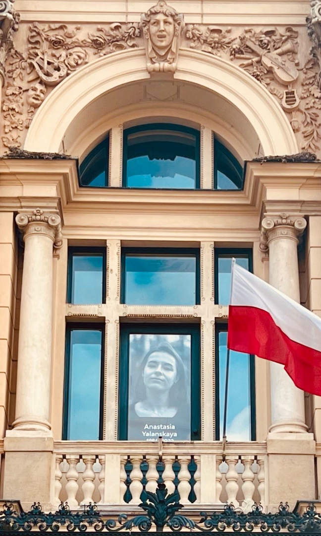 Na zdjęciu fronte okna głównego budynku Teatru. Po lewej i po prawej stronie okna ozdobne kolumny. Nad oknem zdobione rzeźby. W środkowej szybie zdjęcie młodej dziewczyny w kolorze czarno-białym. Po prawej stronie zdjęcia zawieszona polska flaga.