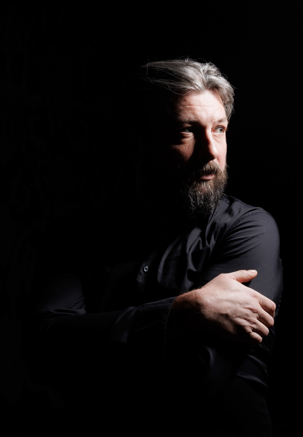 46-letni Krzysztof Wrona w ujęciu portretowym. Mężczyzna ma siwe, zaczesane do tyłu włosy oraz pełną, siwiejącą brodę i wąsy. Ubrany jest w czarną koszulę lub marynarkę. Stoi bokiem do aparatu i spogląda w prawą stronę kadru. Jedną ręką obejmuje drugie ramię, tworząc zamkniętą, skupioną pozę. Twarz i włosy są mocno oświetlone z jednej strony, pozostała część sylwetki całkowicie wtapia się w czarne tło.