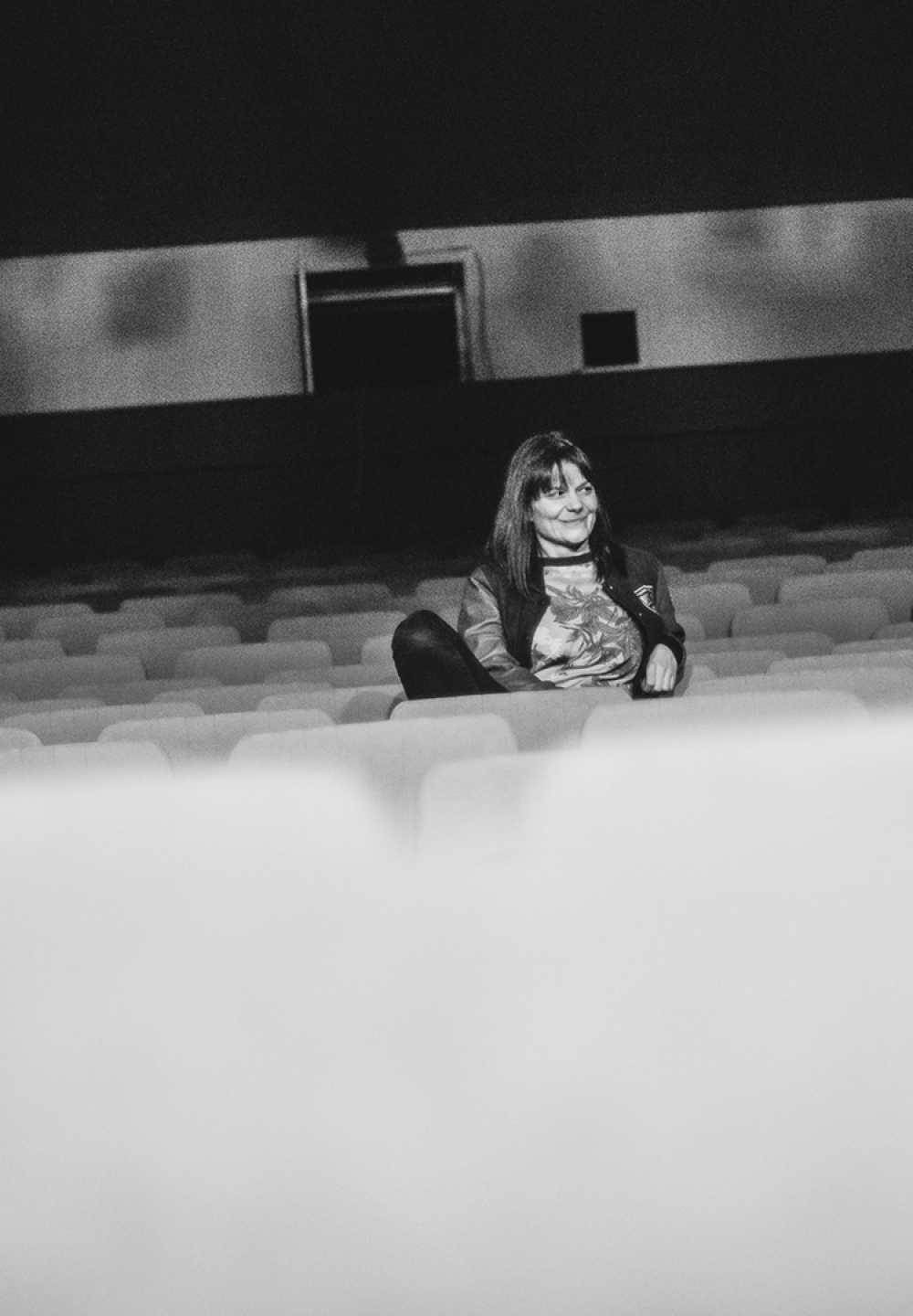 Na zdjęciu reżyserka Magdalena Miklasz. Czarno-biała fotografia. Kobieta siedzi w środku rzędu na widowni. Ma długie, ciemne włosy. Patrzy w prawo. Uśmiecha się. Za nią niewielkie okna z pokoju technicznego.